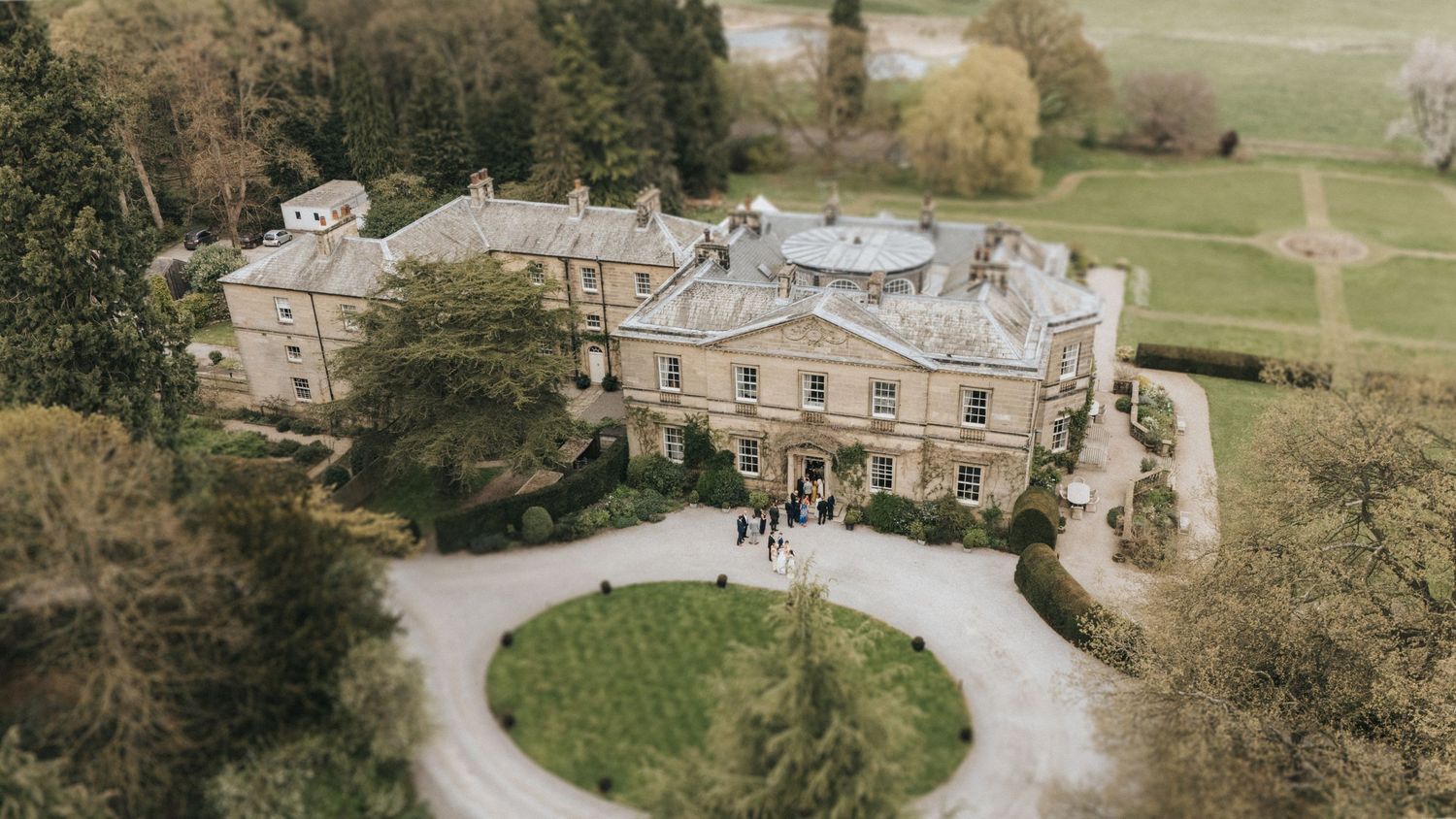 Aerial view of a grand English manor house with circular driveway and manicured grounds surrounded by woodland.