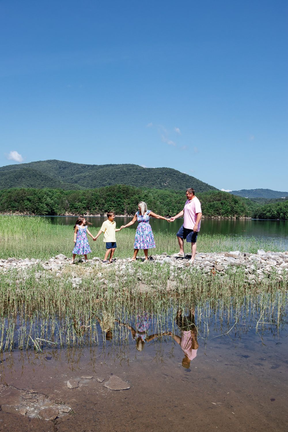 Family of four holds hands while walking across rocks near a lake, with clear skies and mountain views behind them.
