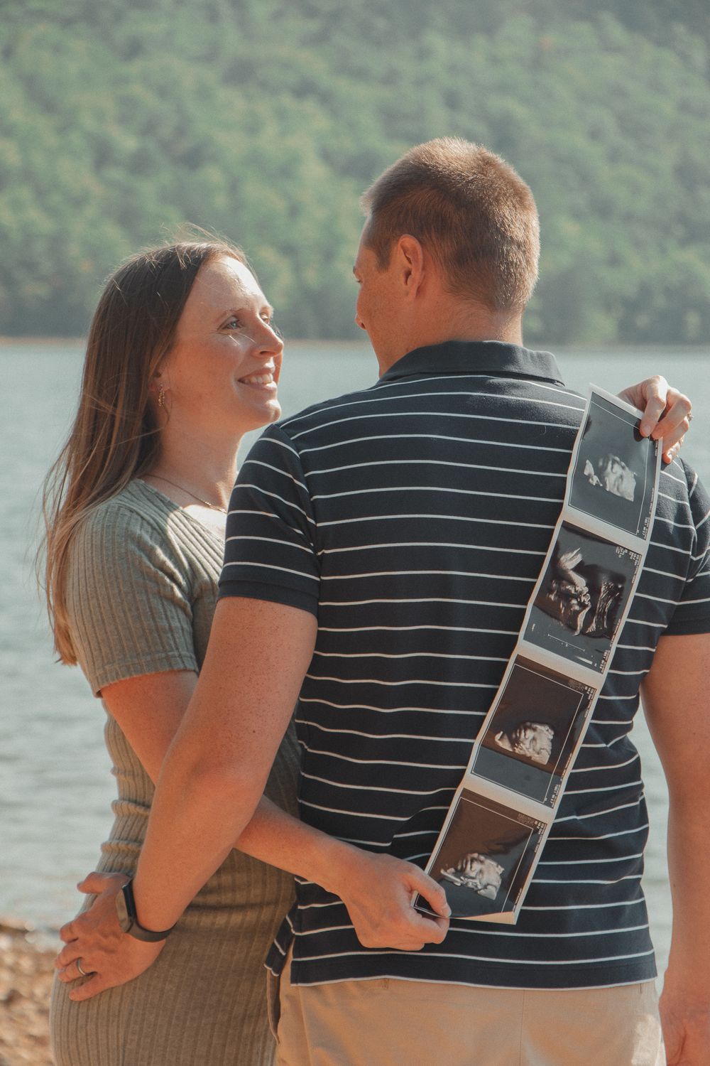Couple smiles at each other while holding ultrasound photos, standing near a lake with trees in the background