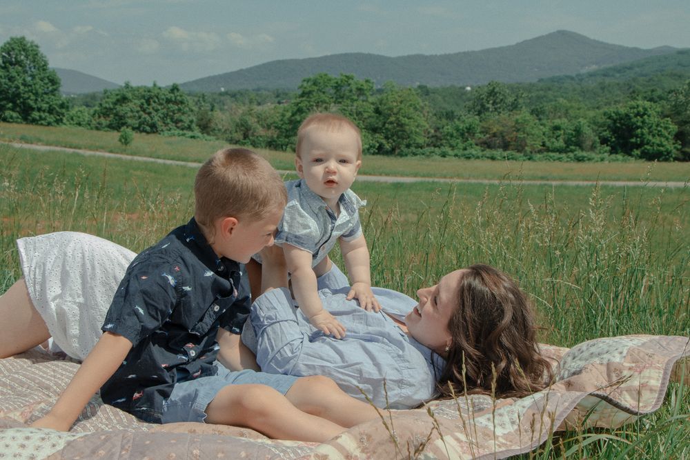 Mom lies on a blanket in a field as her two boys play on top of her, surrounded by grass and mountain views.