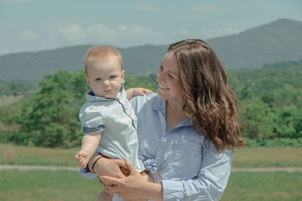 Mom holds baby on her hip, smiling at him, with trees and mountains in the distance under a bright sky