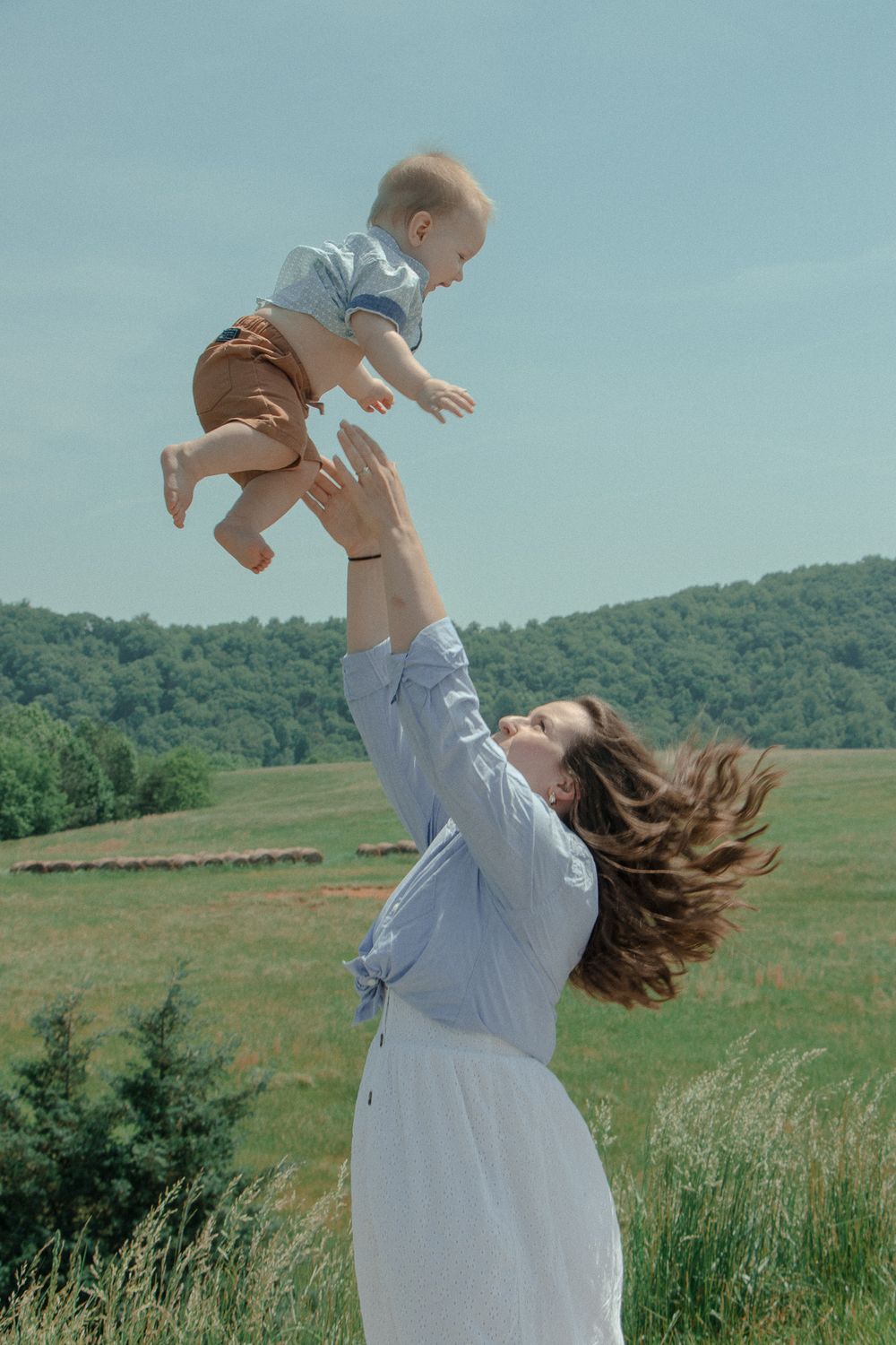 Mom joyfully lifts her baby into the air in a green field, with bright sunshine and blue skies overhead.