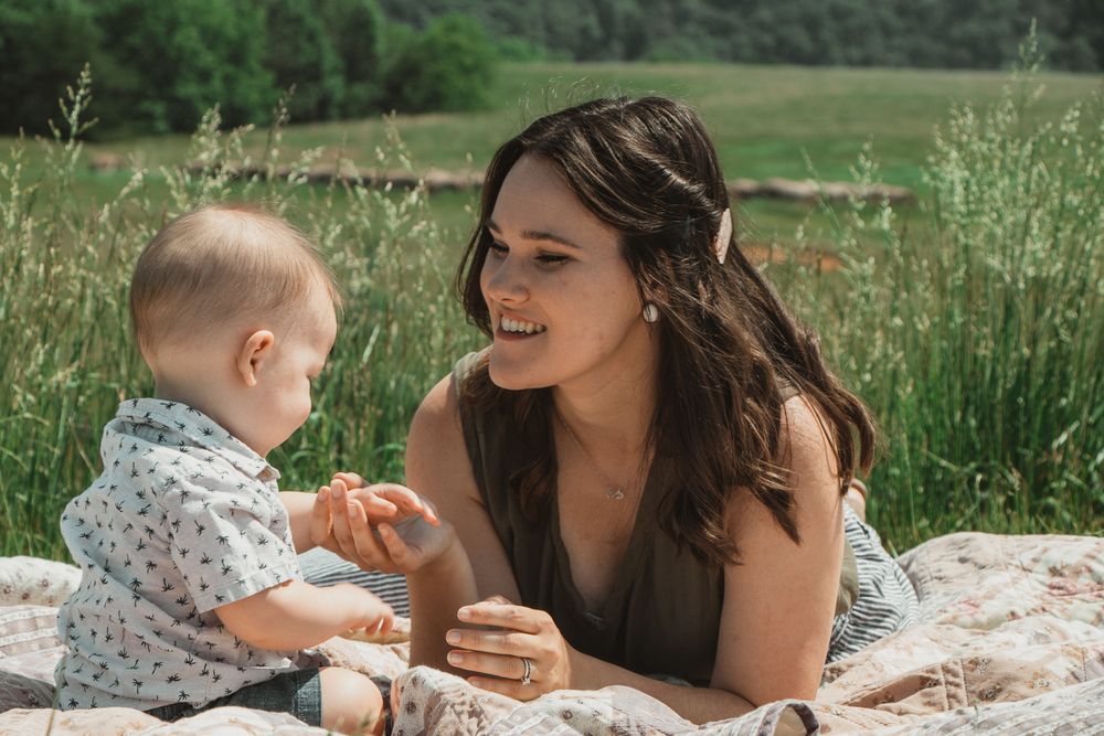 Mom and baby play on a quilt in a grassy field, smiling and holding hands during a sunny day.