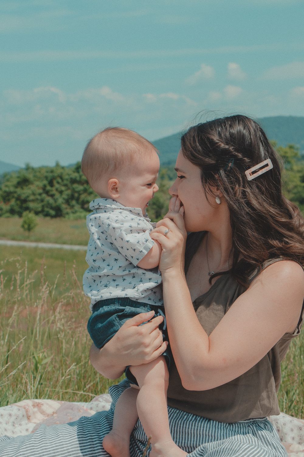 Mom and baby share a joyful moment in a field, touching noses and smiling under a bright blue sky