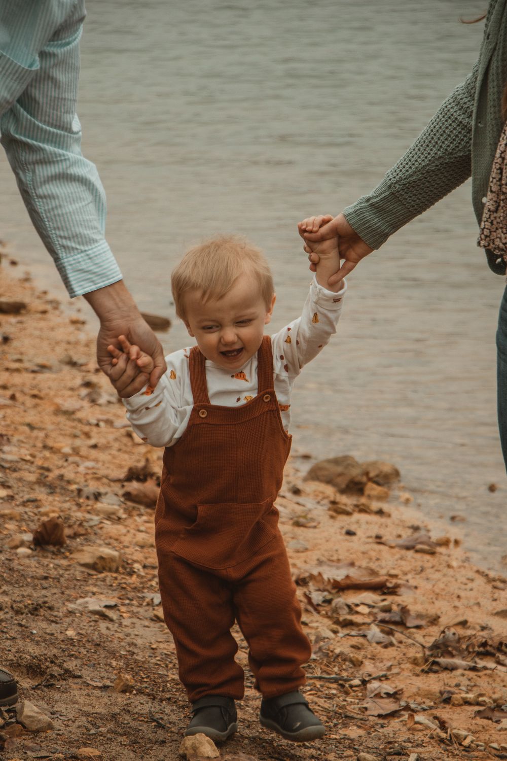 Toddler laughs while holding hands with parents, walking along the shoreline in rust overalls and a printed shirt.