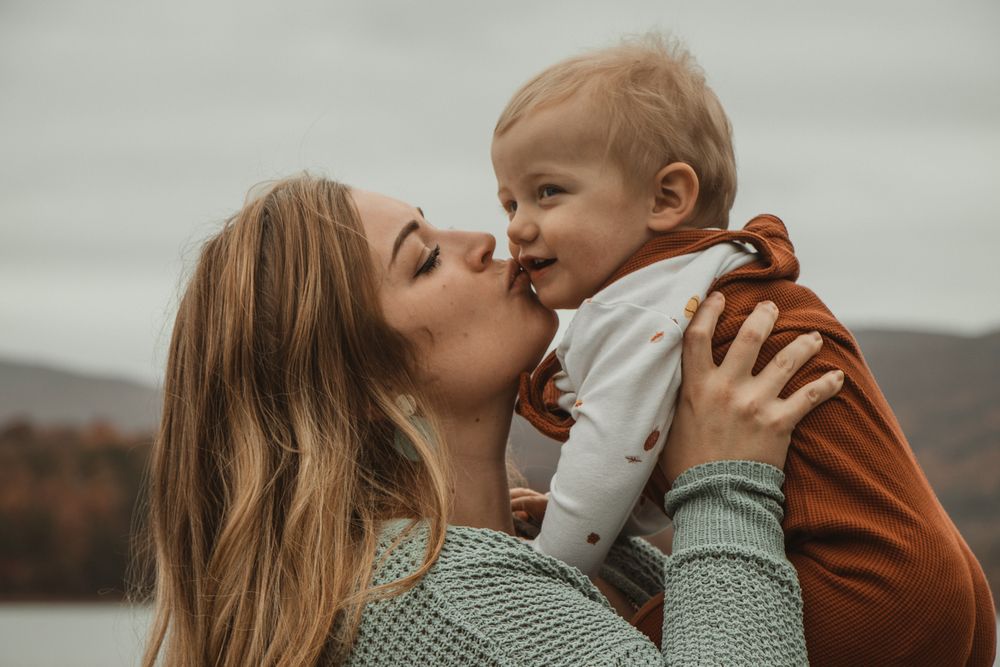 Mom lifts and kisses her smiling toddler while standing near a lake on a cloudy day.