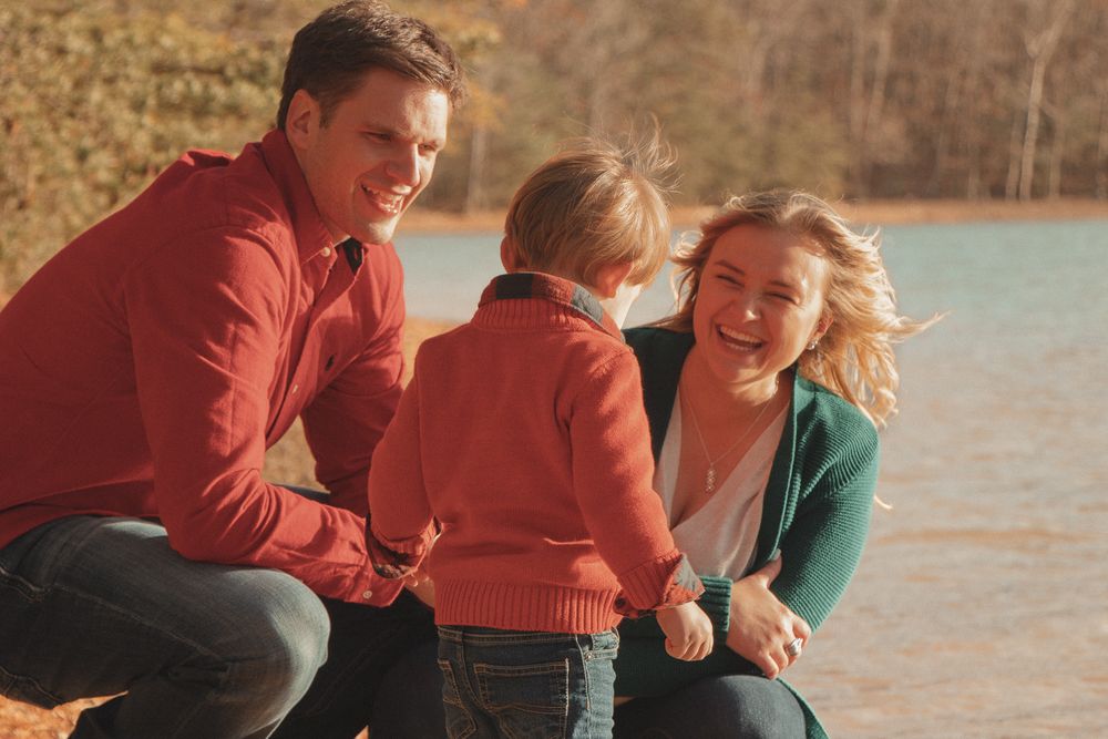 Parents laugh with their toddler at the lakeside, enjoying golden-hour light and candid joy.