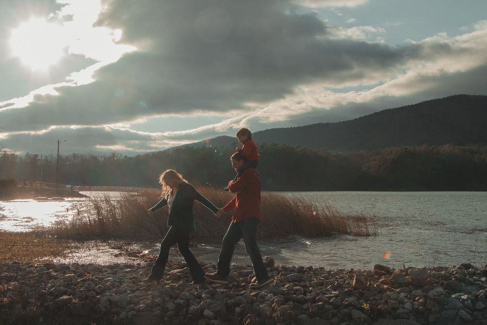 Family walks beside a lake at sunset, with golden light behind them and a child riding on dad's shoulders