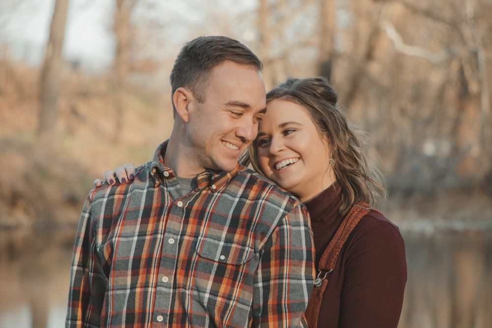 Couple stands close and smiling, leaning into each other with soft golden light and trees behind them.
