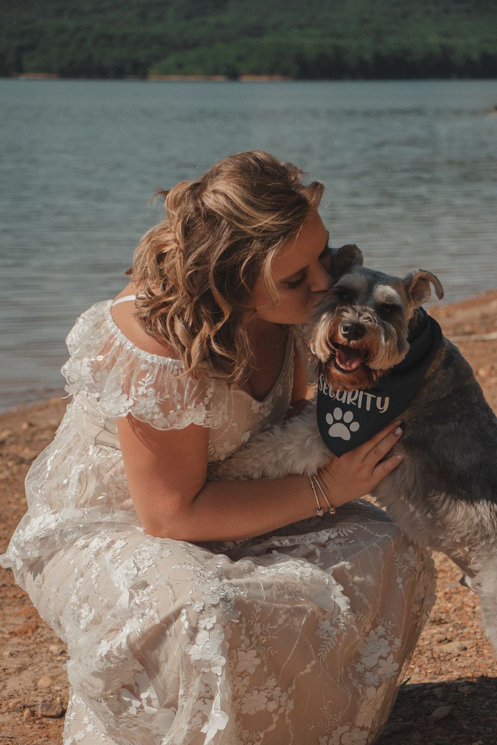 Woman in lace dress kisses her happy dog wearing a "security" bandana, by a lakeshore with forest in the distance.