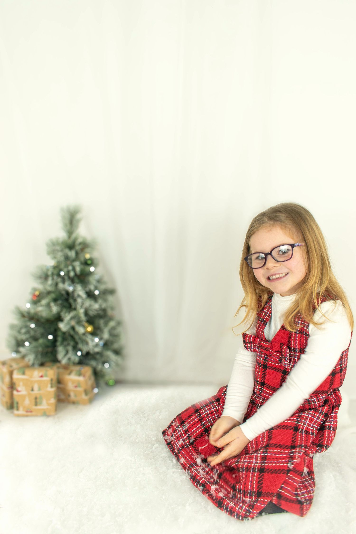Holiday photo session with Christmas tree and plaid dress against white backdrop.