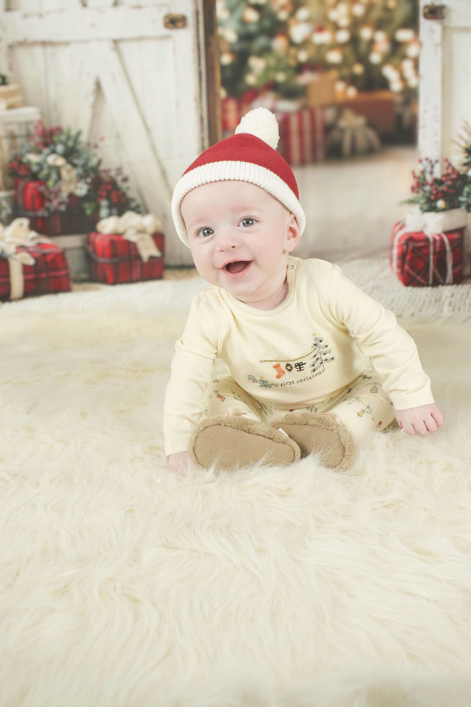 Baby in red hat and cream outfit lying on plush white rug with holiday decorations behind.