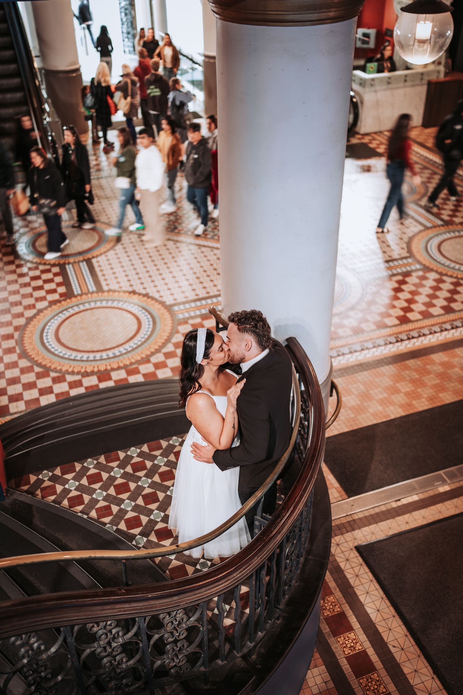 A couple shares an intimate moment on ornate stairs with tiled floors and white columns while people walk below.