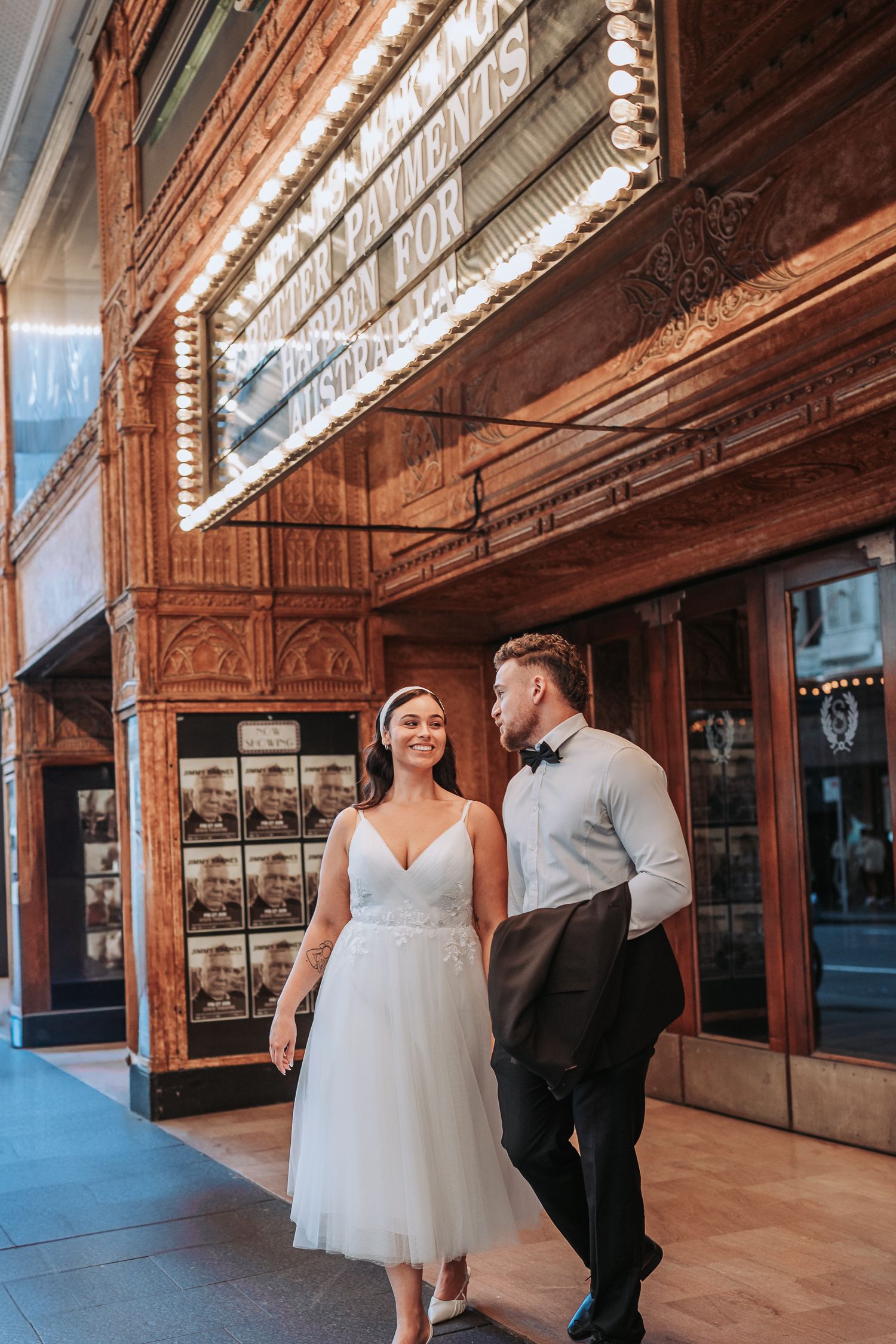 A couple in wedding attire stands together in an elegant venue with wooden trim and large windows lining the walls.
