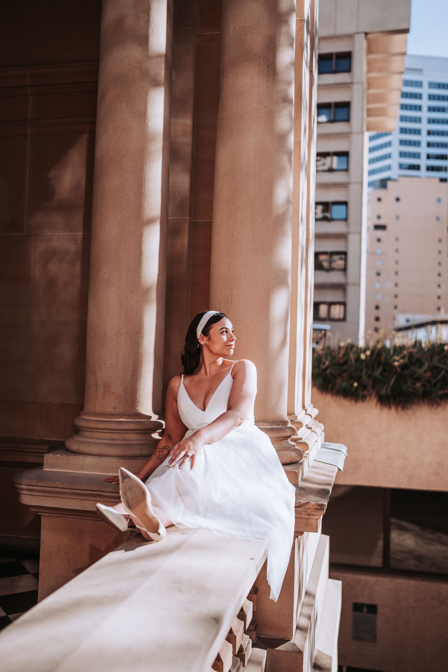 Woman in elegant white wedding dress poses gracefully on a balcony with urban architecture in the background.