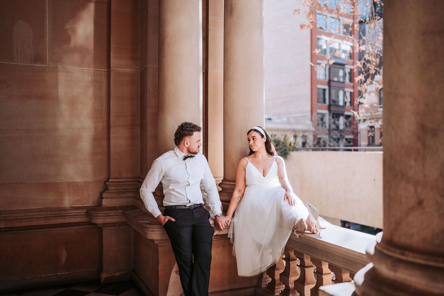 A wedding couple shares an intimate moment in a grand hallway with elegant stone columns and warm lighting.