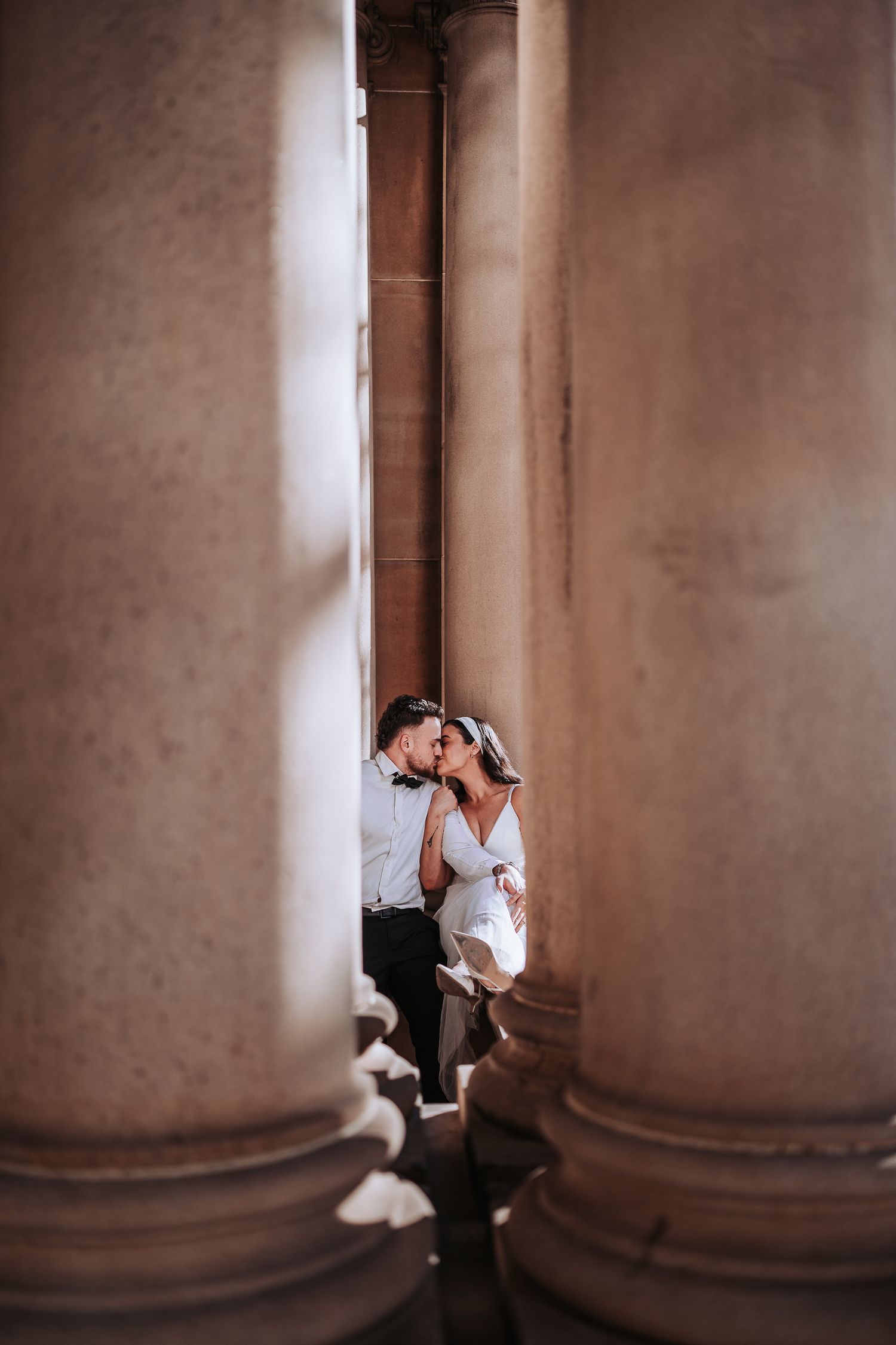 A couple embraces between large stone columns, creating a romantic and intimate architectural composition.