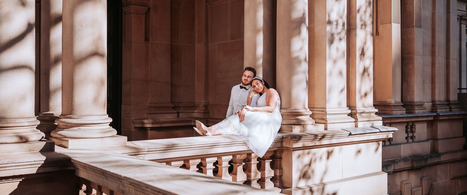 A couple in wedding attire sits together on an elegant stone balcony surrounded by classical marble columns.