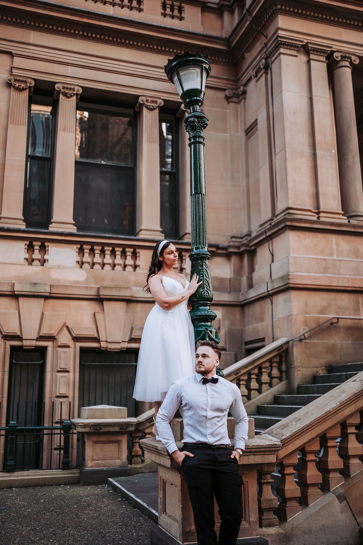 Romantic couple in formal attire pose elegantly on vintage stone staircase with ornate lamp post at historic building.