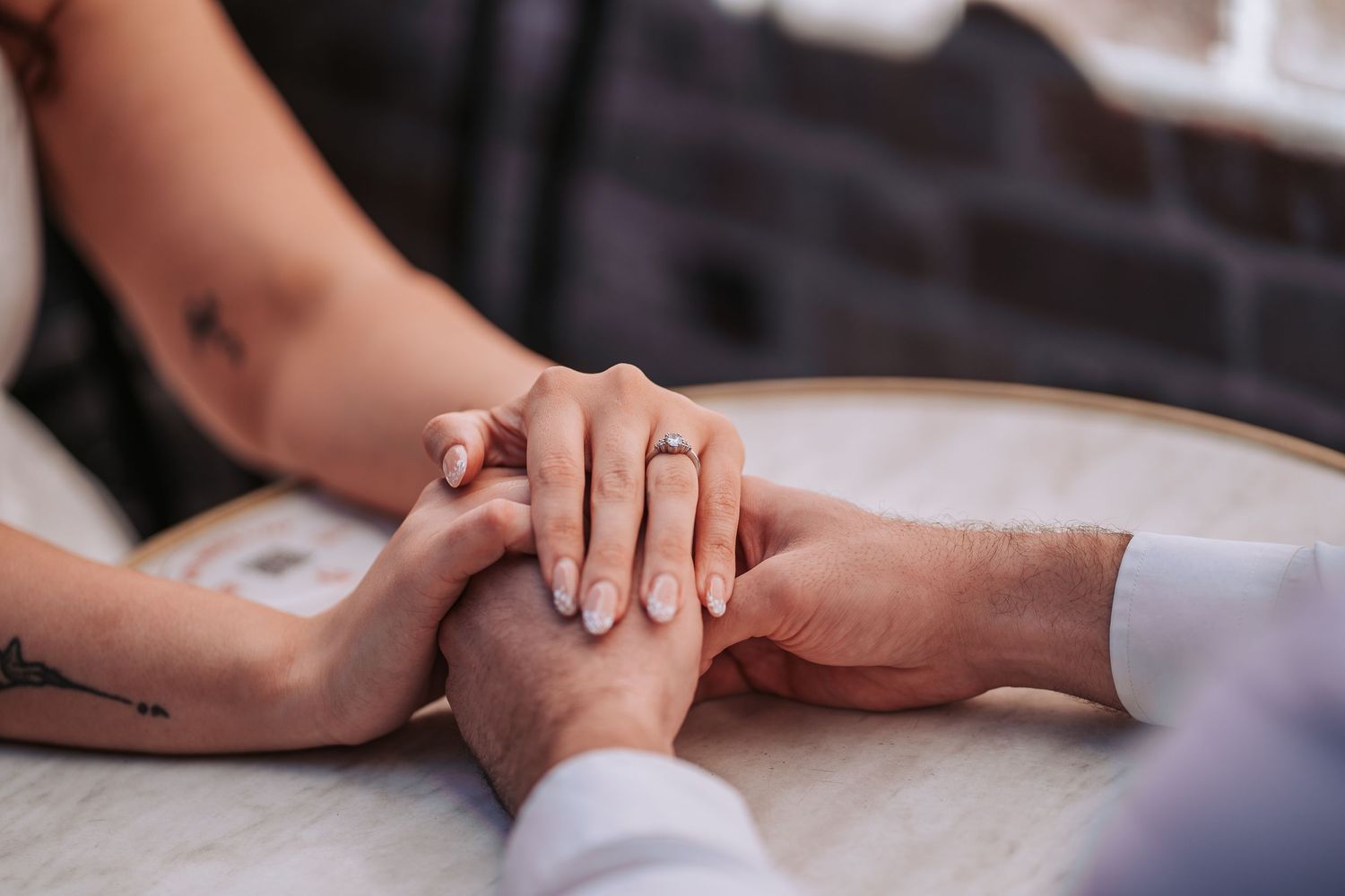 Two hands tenderly clasped together on a white tabletop, one with visible tattoos and an engagement ring.