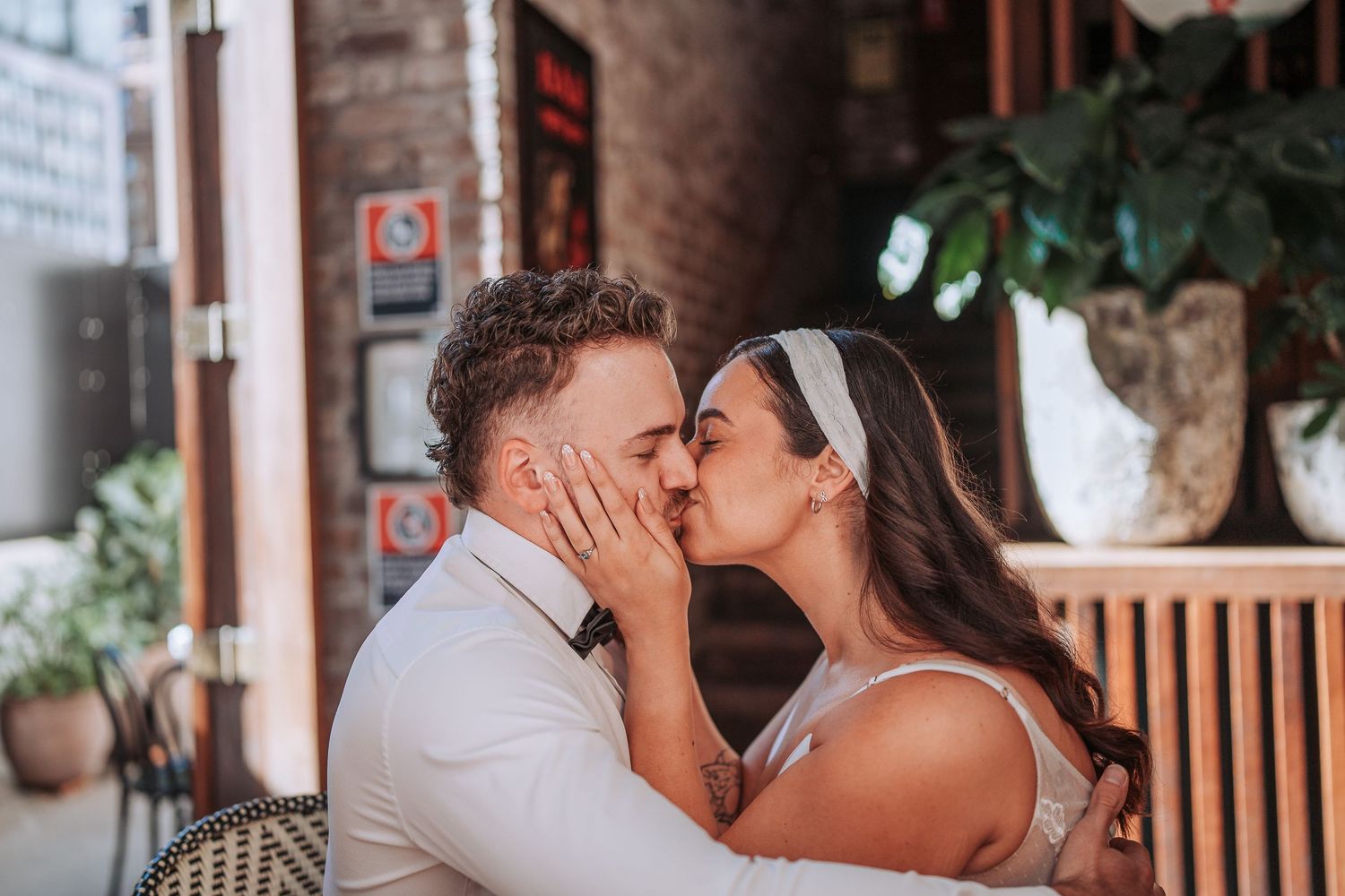 A romantic couple in wedding attire share an intimate moment on a porch with greenery and decor in the background.