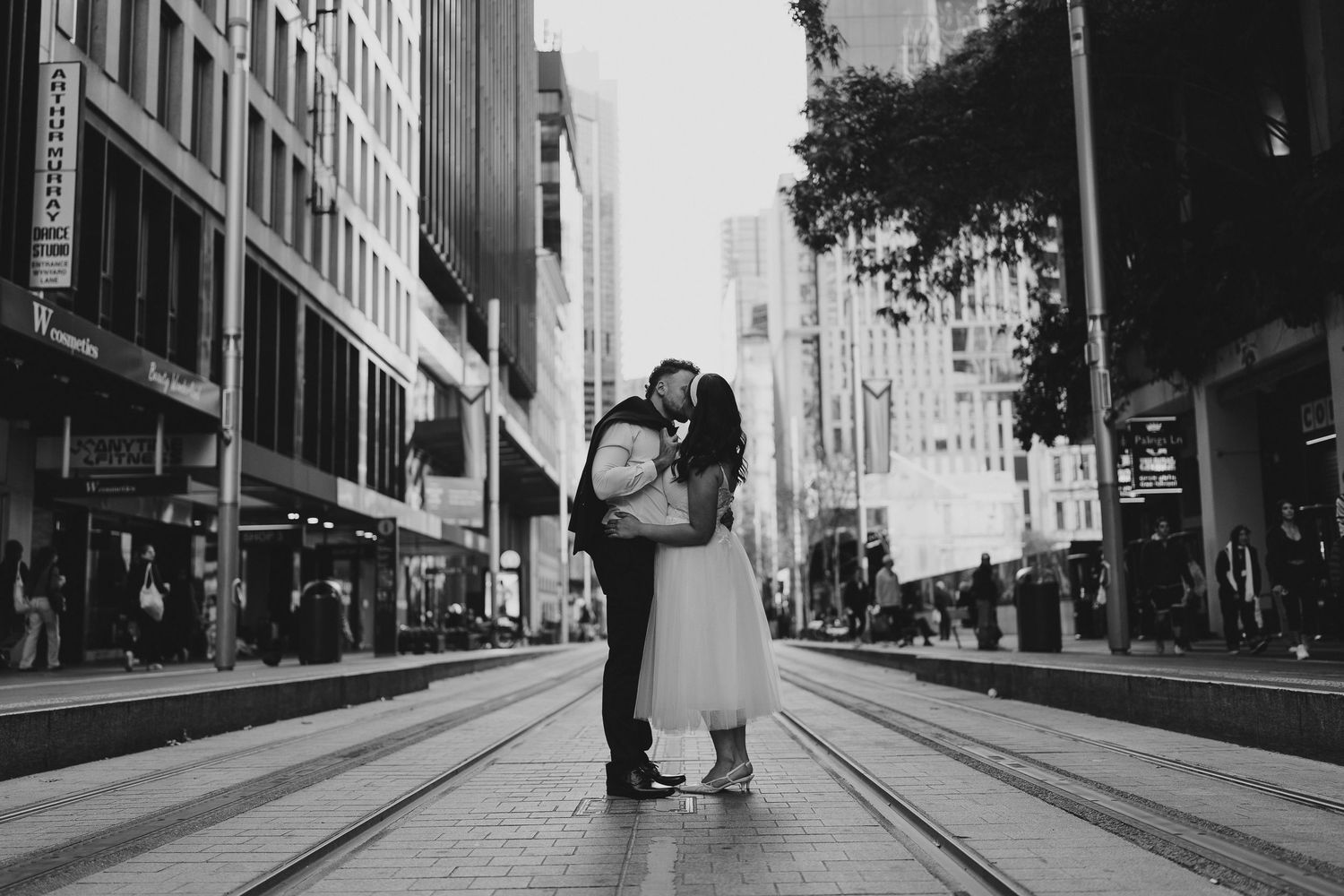 Two people stand on an empty city street lined with tall buildings captured in black and white photography.
