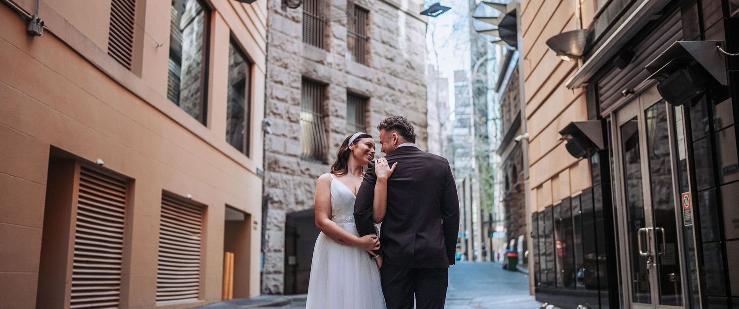 A couple in formal attire walk down a narrow cobblestone alley between historic stone buildings.