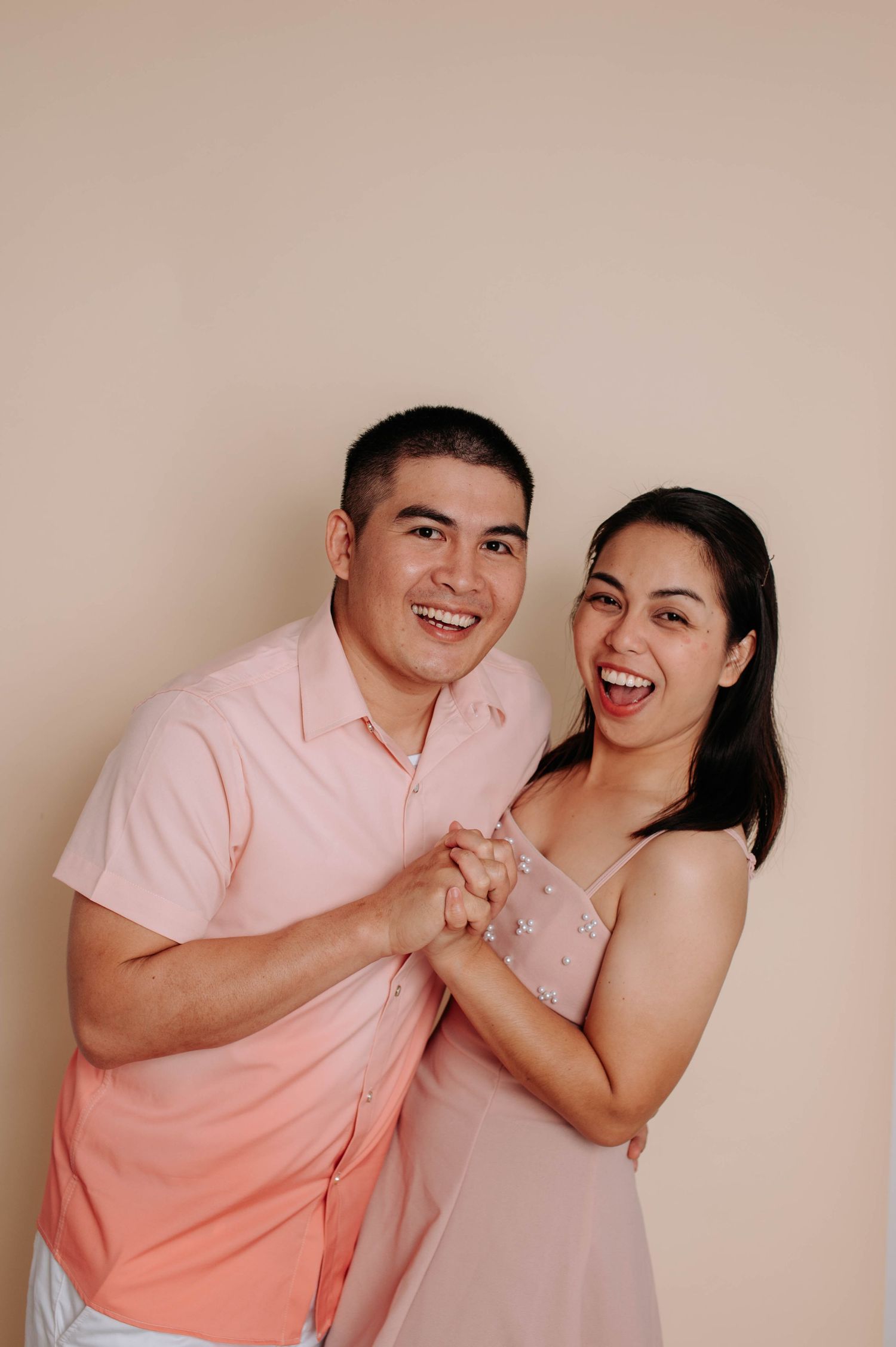Studio portrait of an expectant couple in matching light-colored outfits sitting together on a beige couch.