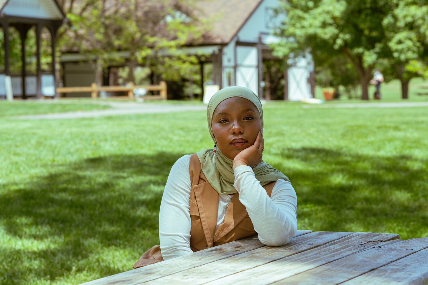 Young Black woman in hijab sits thoughtfully at outdoor wooden table on green lawn near building.