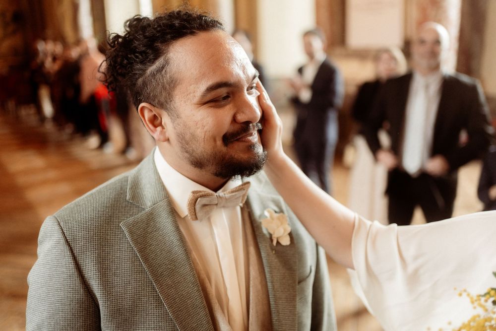 A groom in a light gray suit and bow tie smiles joyfully during an emotional moment at his wedding ceremony.
