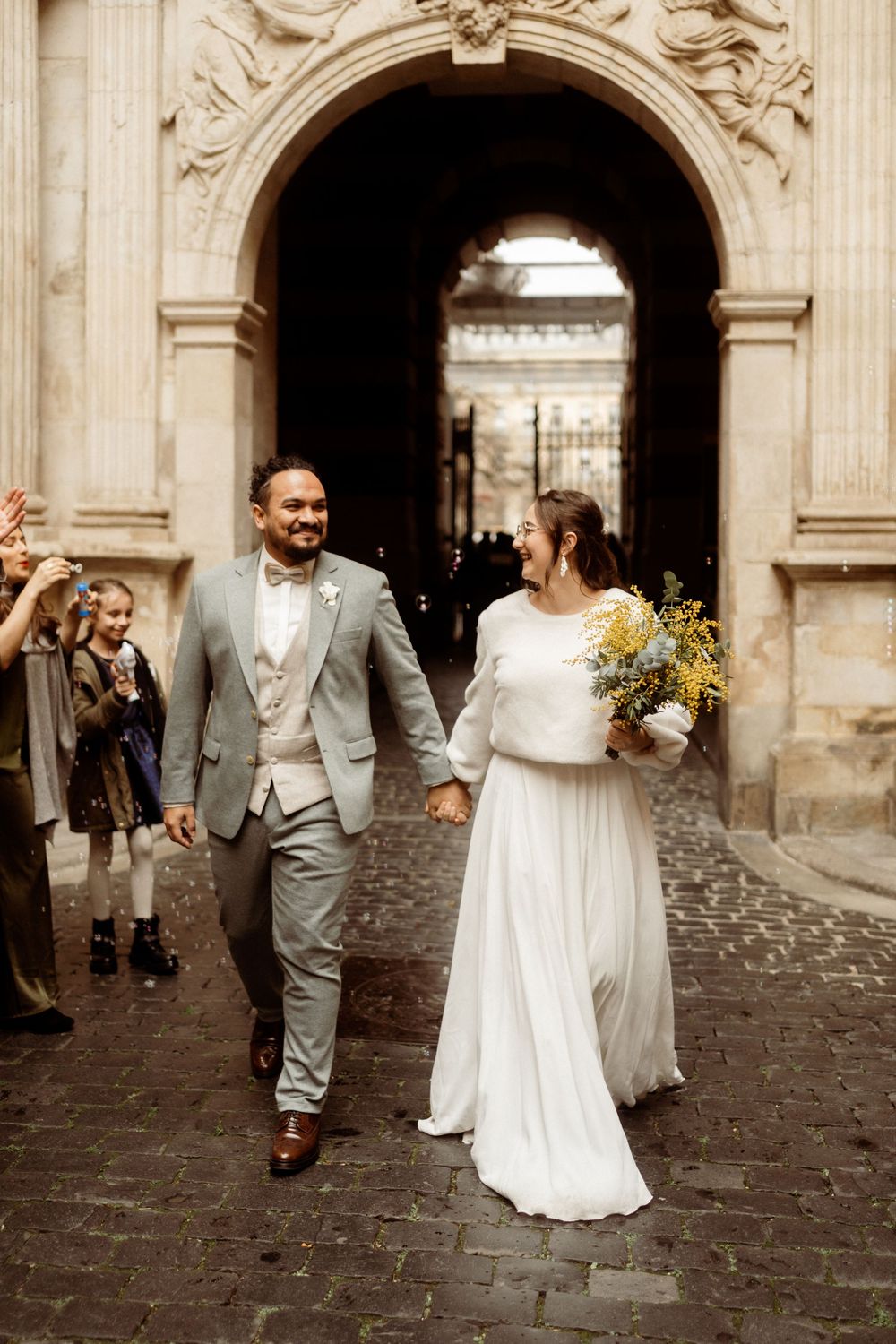 A wedding couple walks through an ornate stone archway on a cobblestone path, with the bride holding yellow flowers.