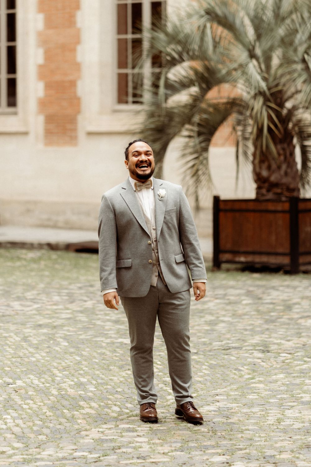 A person in a gray suit stands outdoors on a cobblestone courtyard with palm trees and brick buildings in the background.