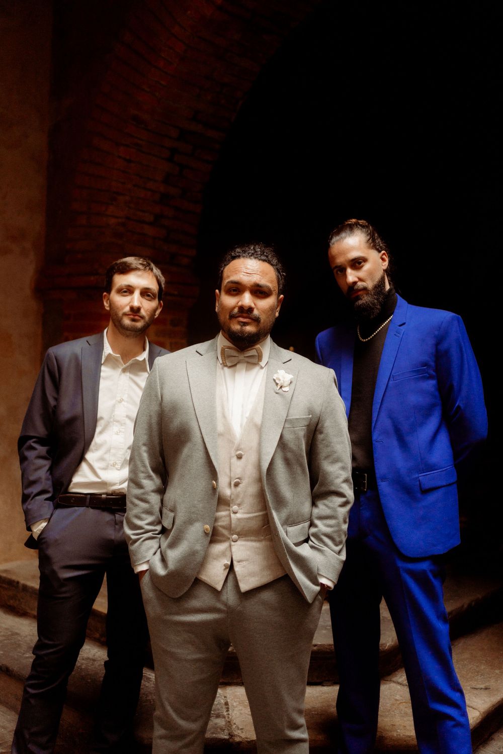 Three groomsmen in formal attire pose in front of a brick archway during a wedding photoshoot.