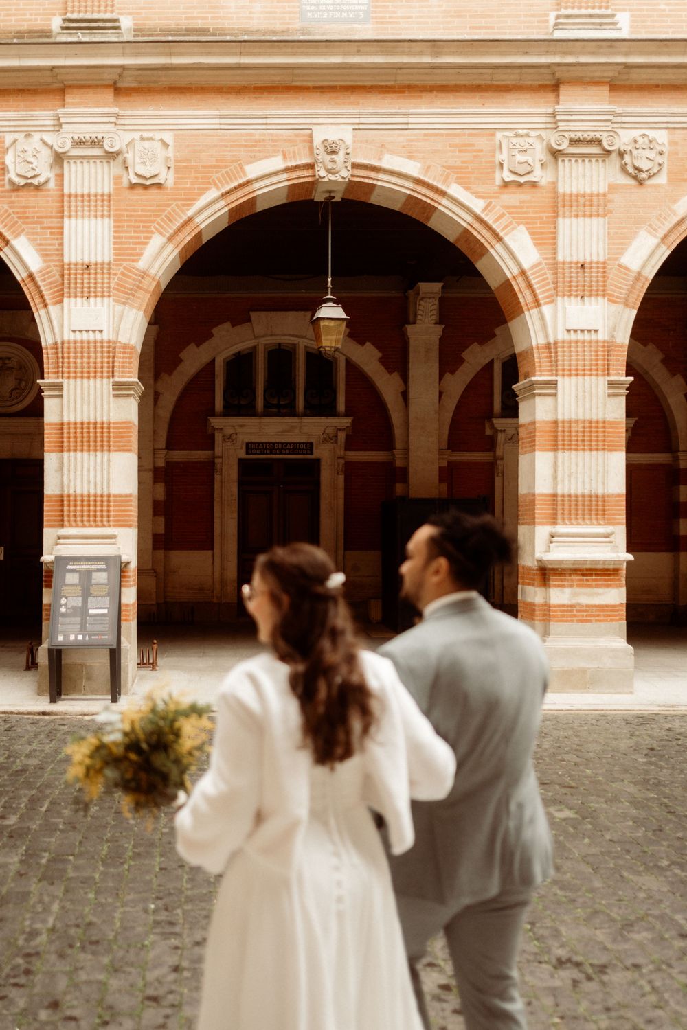 A couple in elegant wedding attire viewed from behind stands beneath ornate terracotta arches of a historic building.