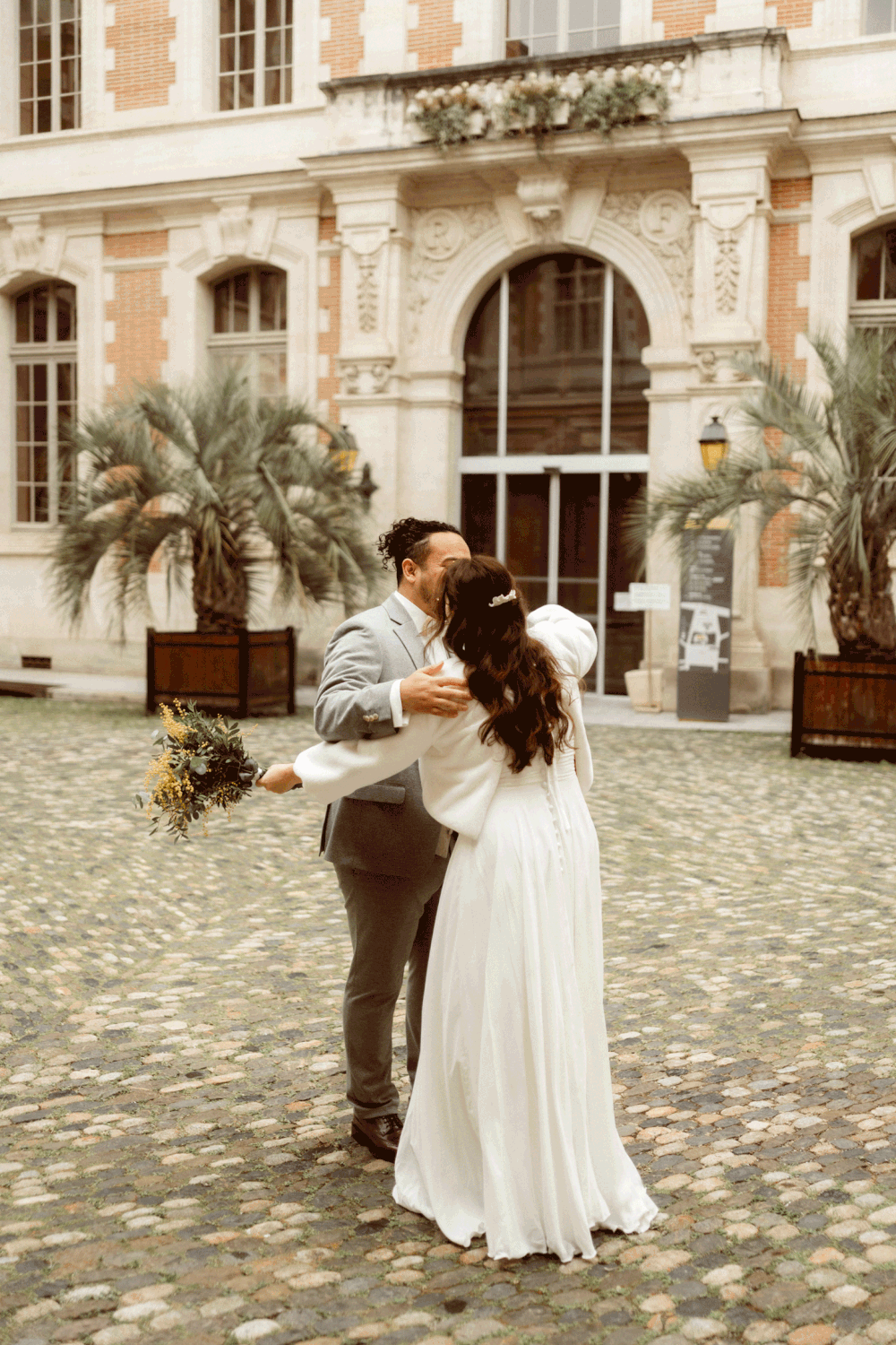 A couple dancing together in front of a historic European building with palm trees and cobblestone courtyard.