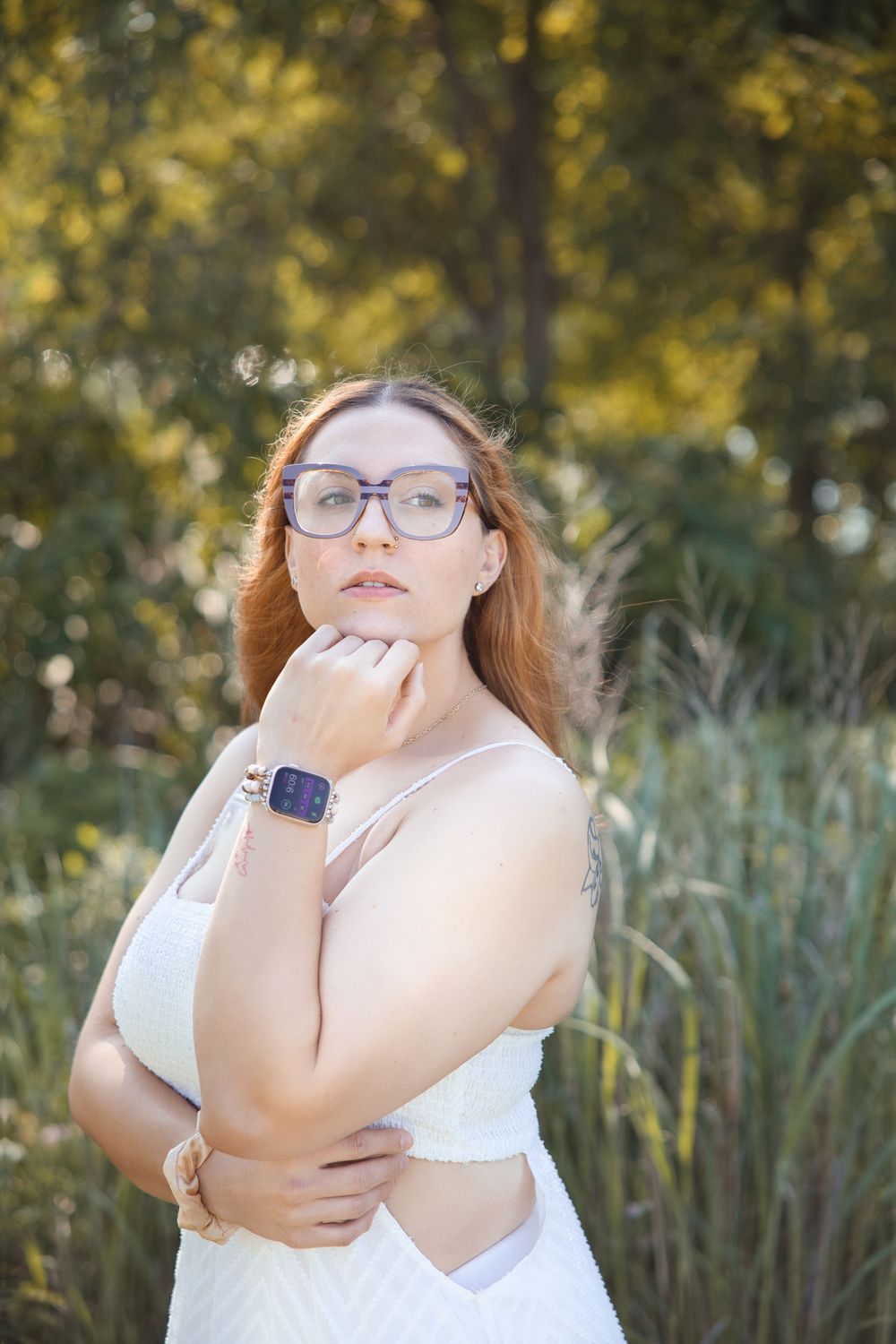 A person with glasses and red hair poses outdoors near tall grass in natural autumn lighting.