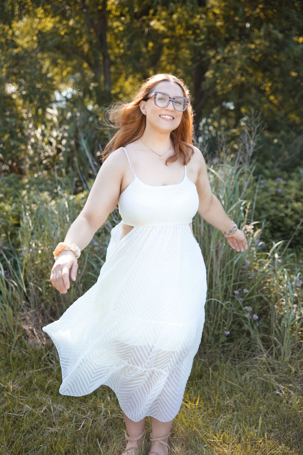 A joyful figure in glasses and a white summer dress twirls outdoors in warm sunlight among tall grasses.