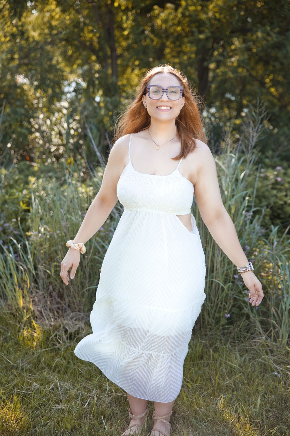 A person in a flowing white summer dress twirls playfully in a sunlit meadow with tall grass and greenery.