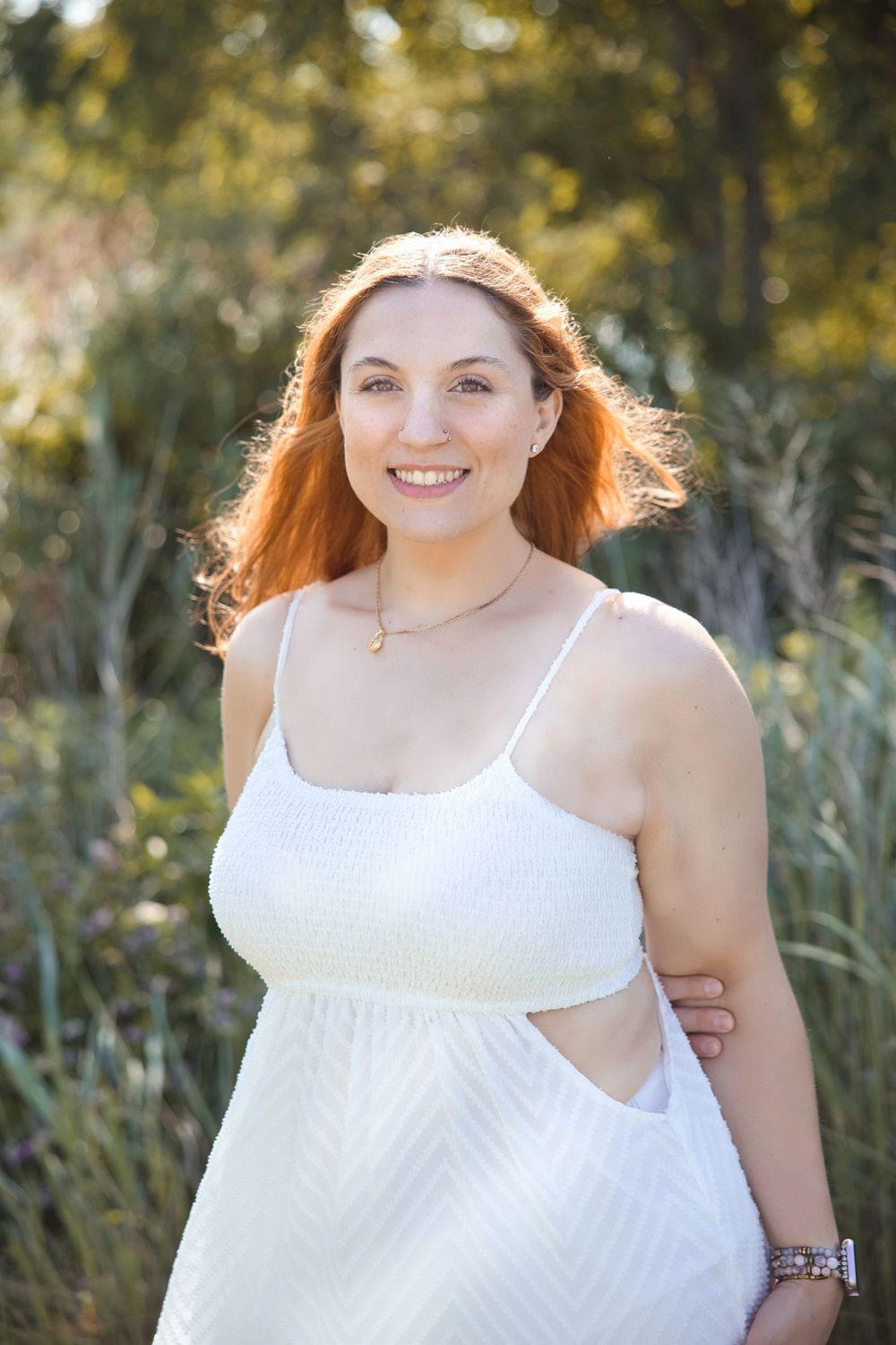 A person wearing a flowing white sundress poses outdoors in a natural setting with sun-lit foliage in the background.