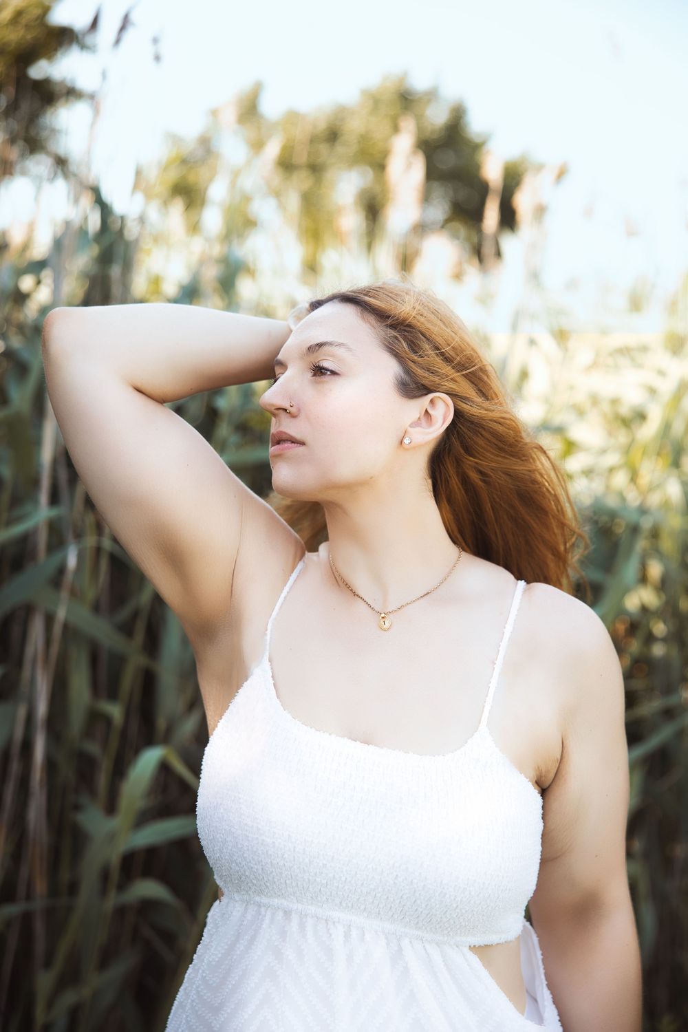 Person in white spaghetti strap dress poses with eyes closed against natural outdoor setting.