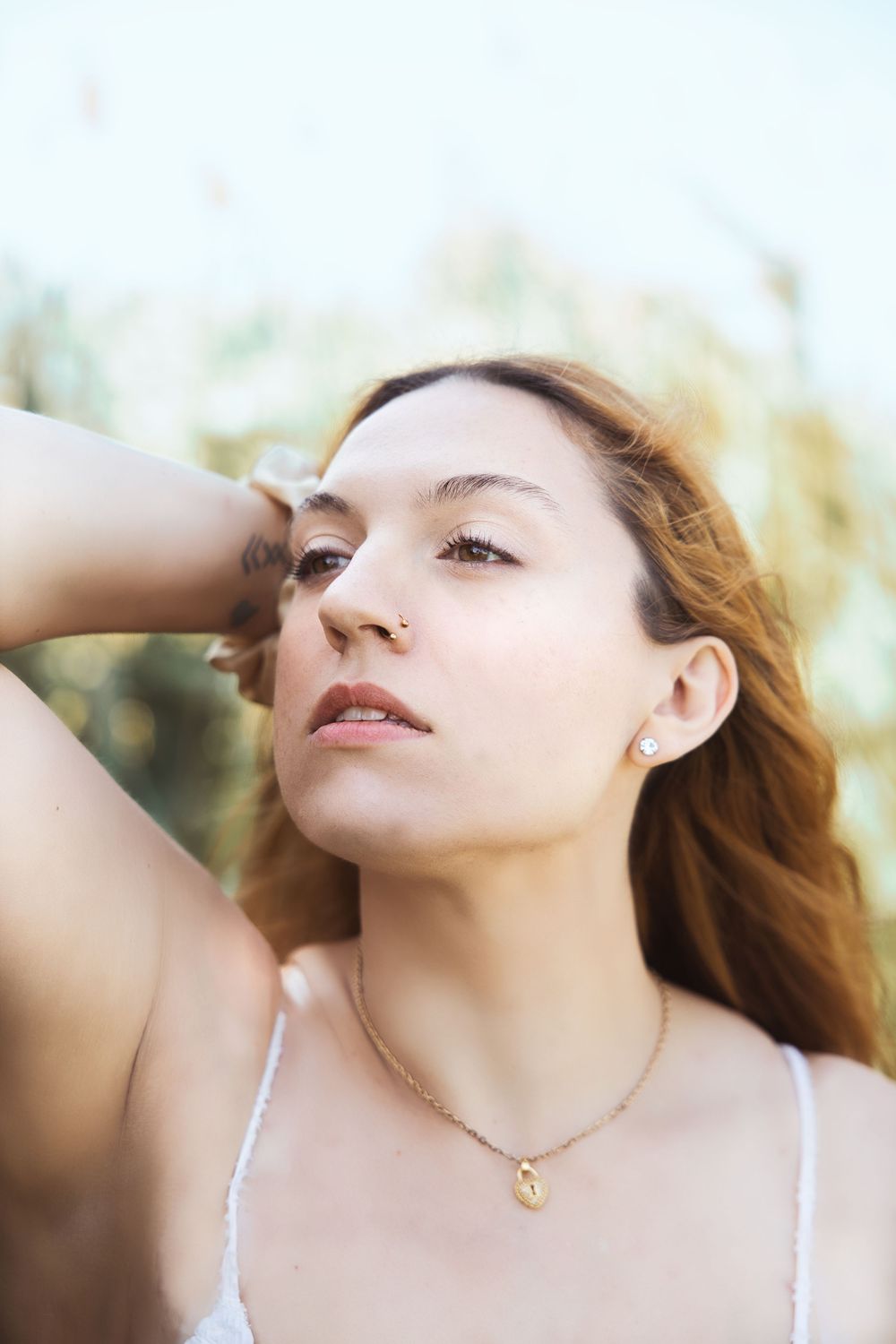 Natural light portrait of a person with flowing hair wearing a white top and delicate necklace against a soft blurred background.