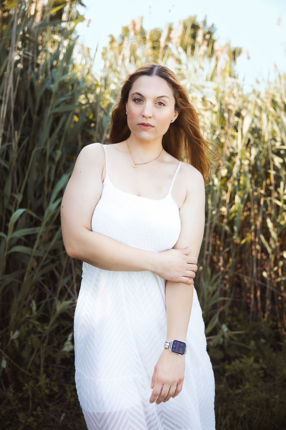 Person in a white summer dress poses against tall reeds in golden sunlight, wearing a smartwatch.