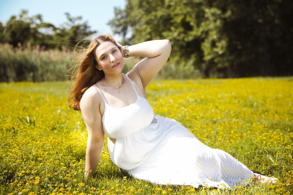 A woman in a flowing white dress reclines in a sun-drenched meadow filled with bright yellow wildflowers.