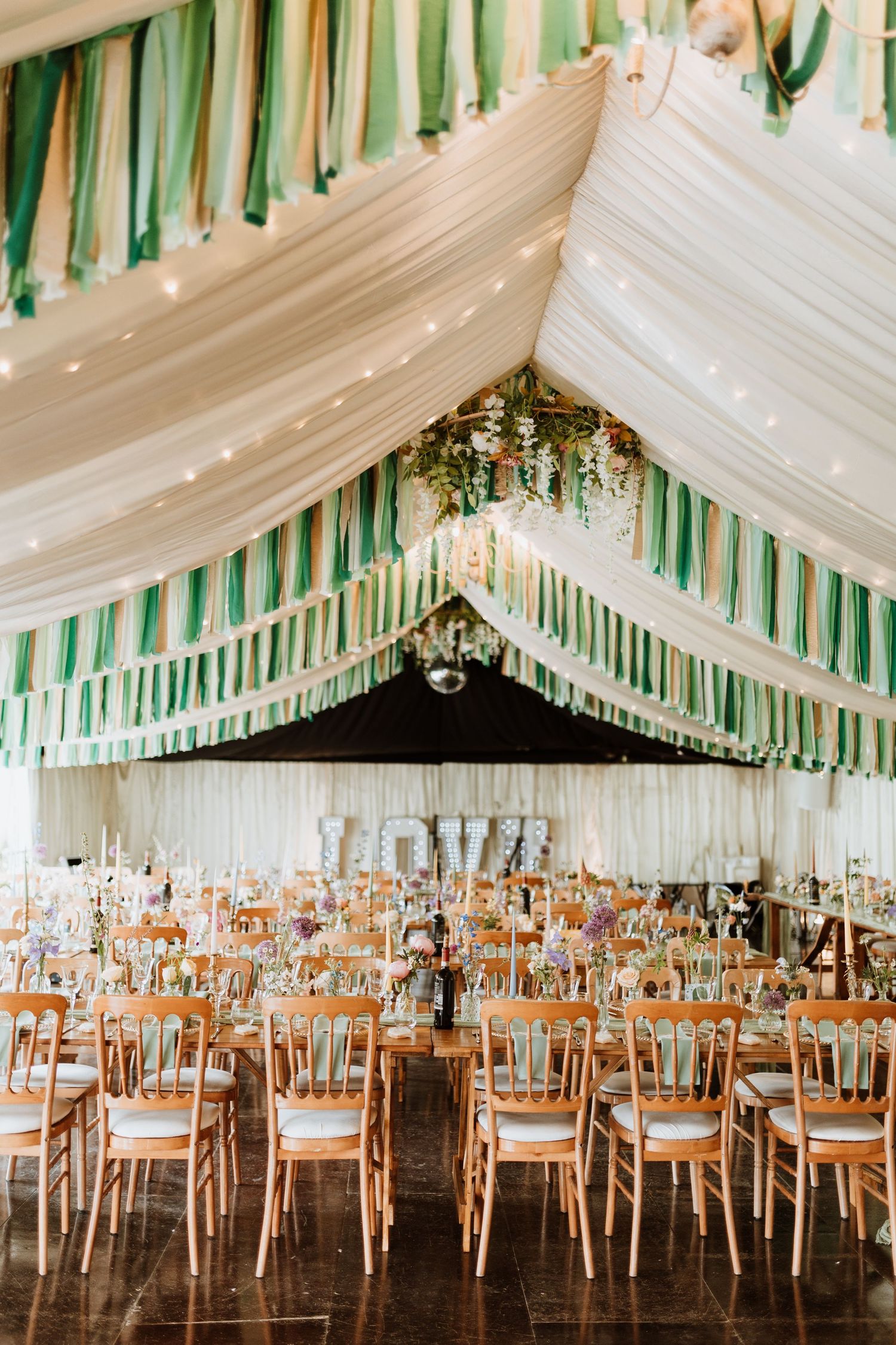 Elegant wedding reception venue with white draped ceiling, green accents, and rows of wooden chairs and tables.
