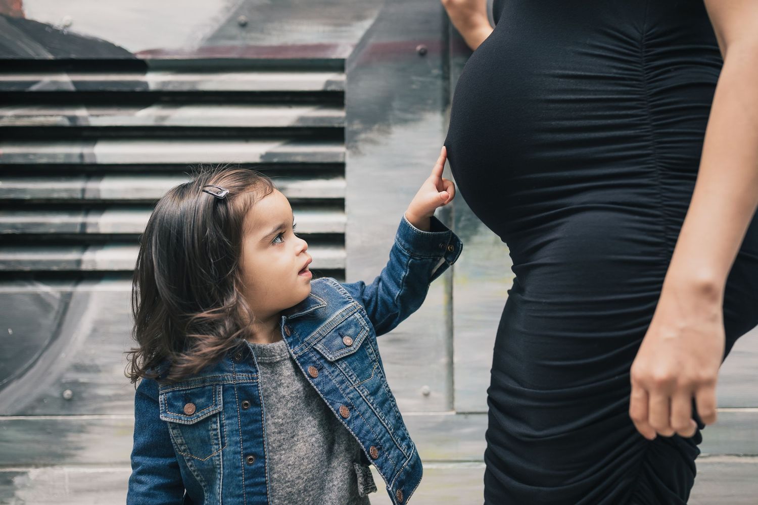 Small child in denim jacket reaches up to a pregnant woman wearing black dress standing near steps.