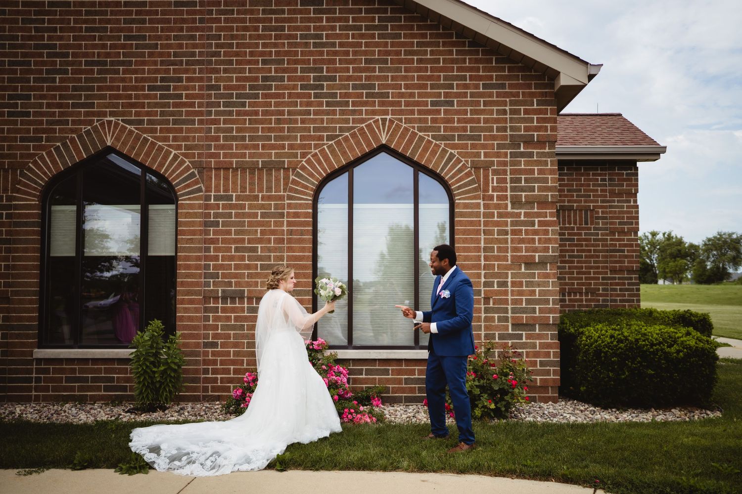 A couple in wedding attire stand before an arched brick church window surrounded by blooming pink roses.