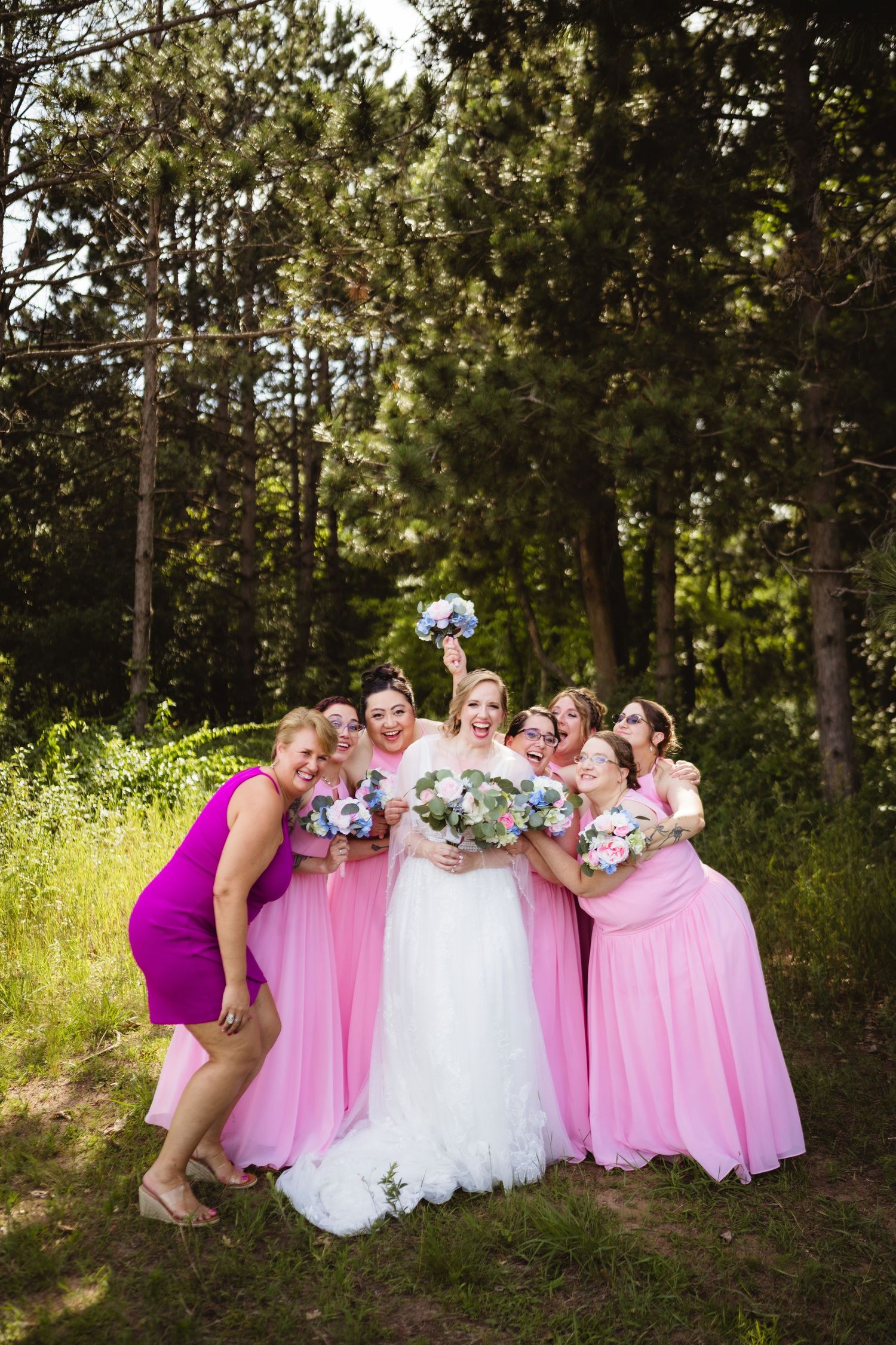 A bridal party in pink dresses huddles together in a playful pose outdoors among pine trees.