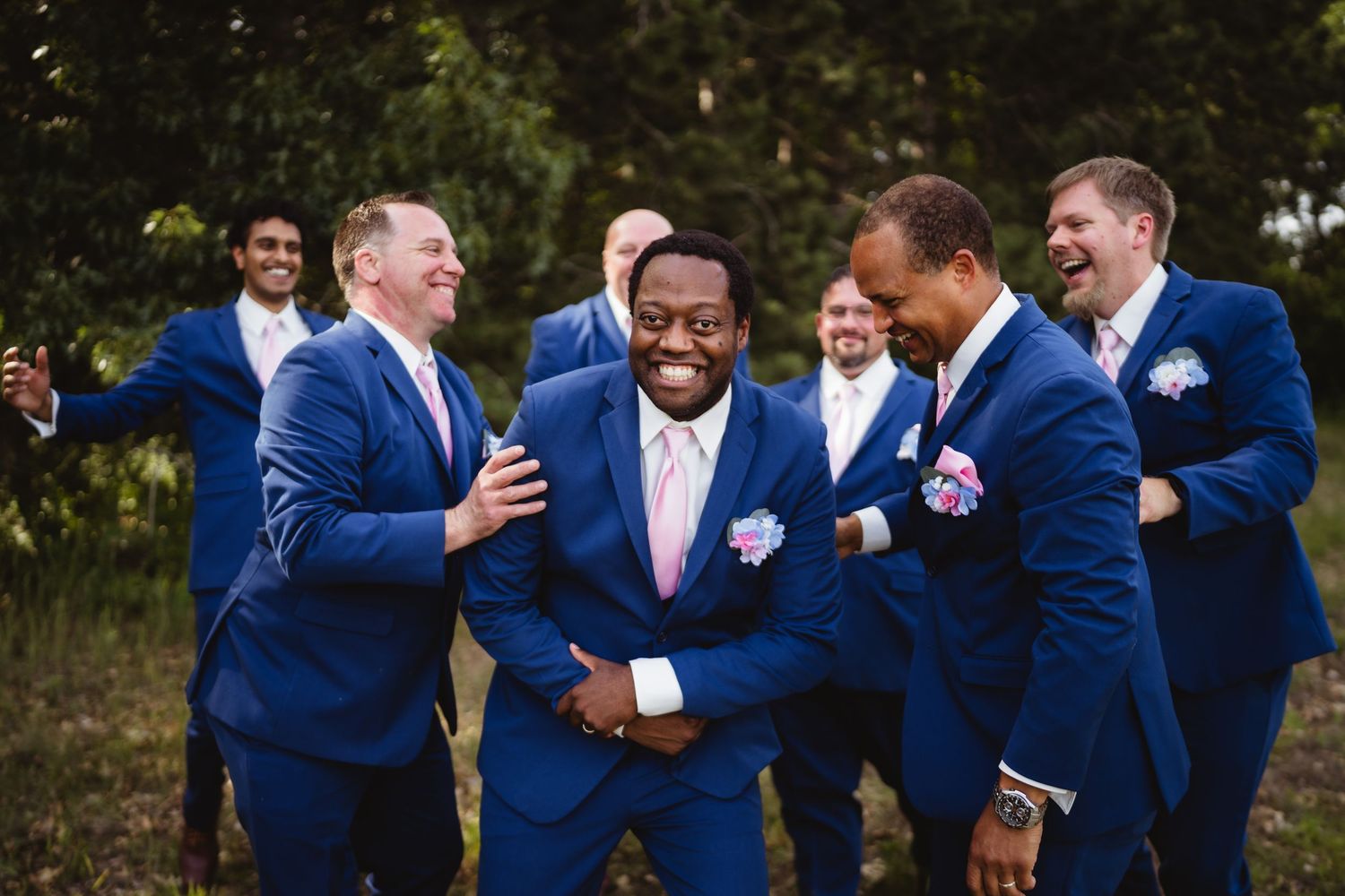 Groomsmen in navy blue suits share candid laughing moments outdoors during wedding photos.