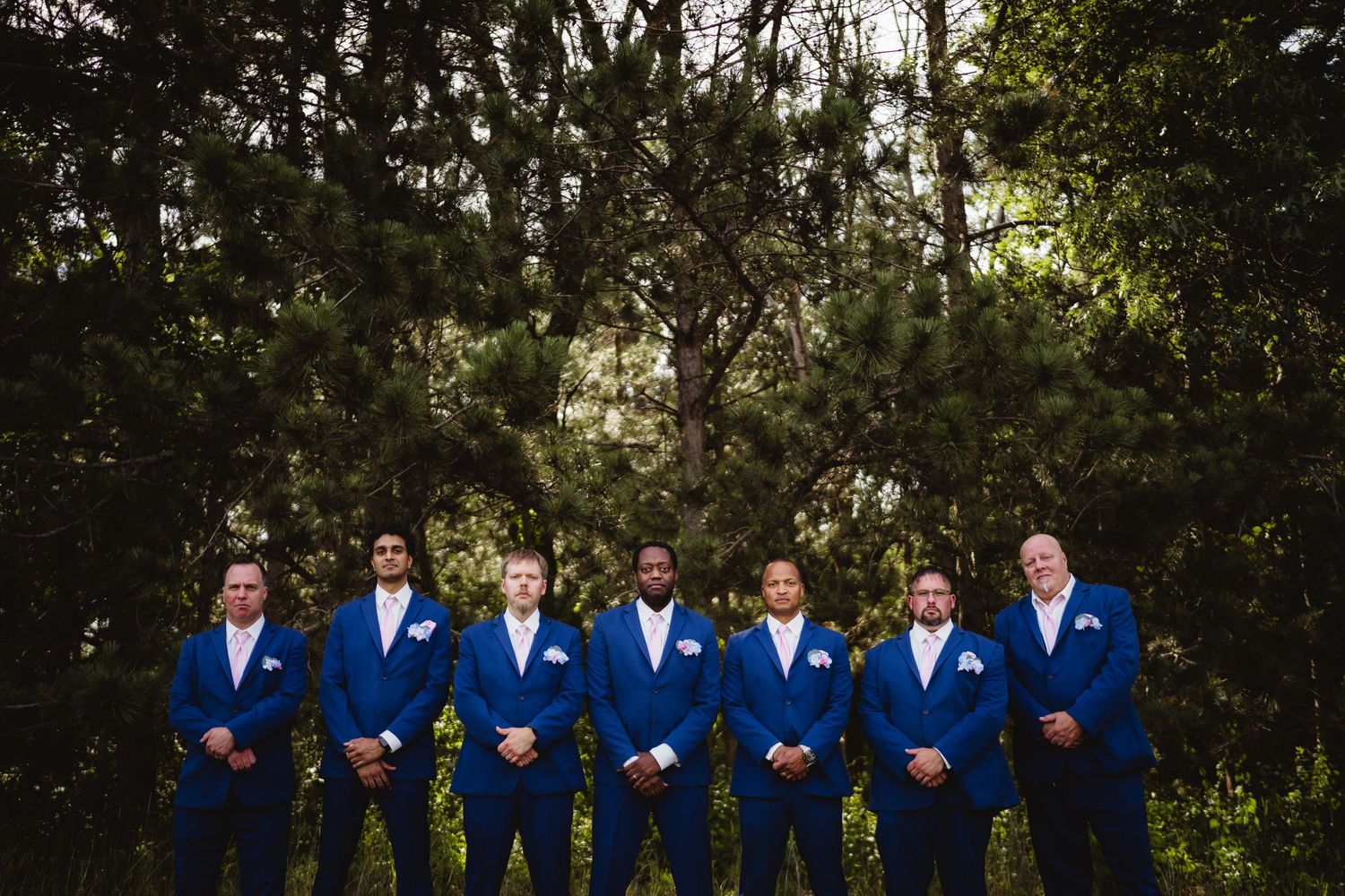 Wedding party wearing coordinated royal blue suits and white boutonnieres pose together in front of wooded backdrop.