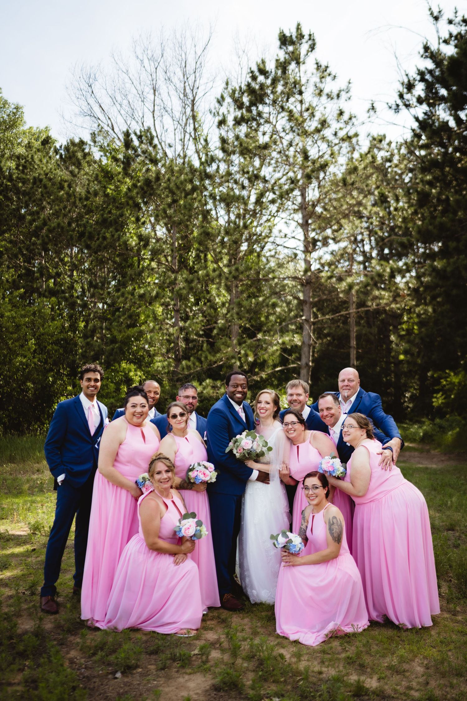 Wedding party in navy blue suits and pink bridesmaid dresses pose together outdoors among pine trees.